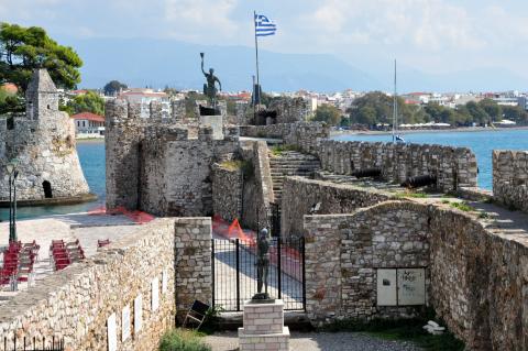 Το άγαλμα του Ανεμογιάννη δίπλα στη ελληνική σημαία και το άγαλμα του Θερβάντες στον βραχίονα του λιμανιού ναυπακτοσ αξιοθεατα